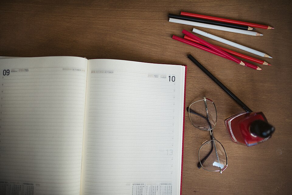 Open planner on a wooden desk beside pencils, glasses and writing supplies